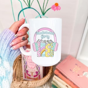 以下が含まれることがあります： ピンクと青のデザインで「Audiobook Girly」の文字が入った白いセラミックマグ。マグには、パステルカラーのヘッドフォンと本のグラフィックが描かれています。背景には本の山とバスケットがあります。