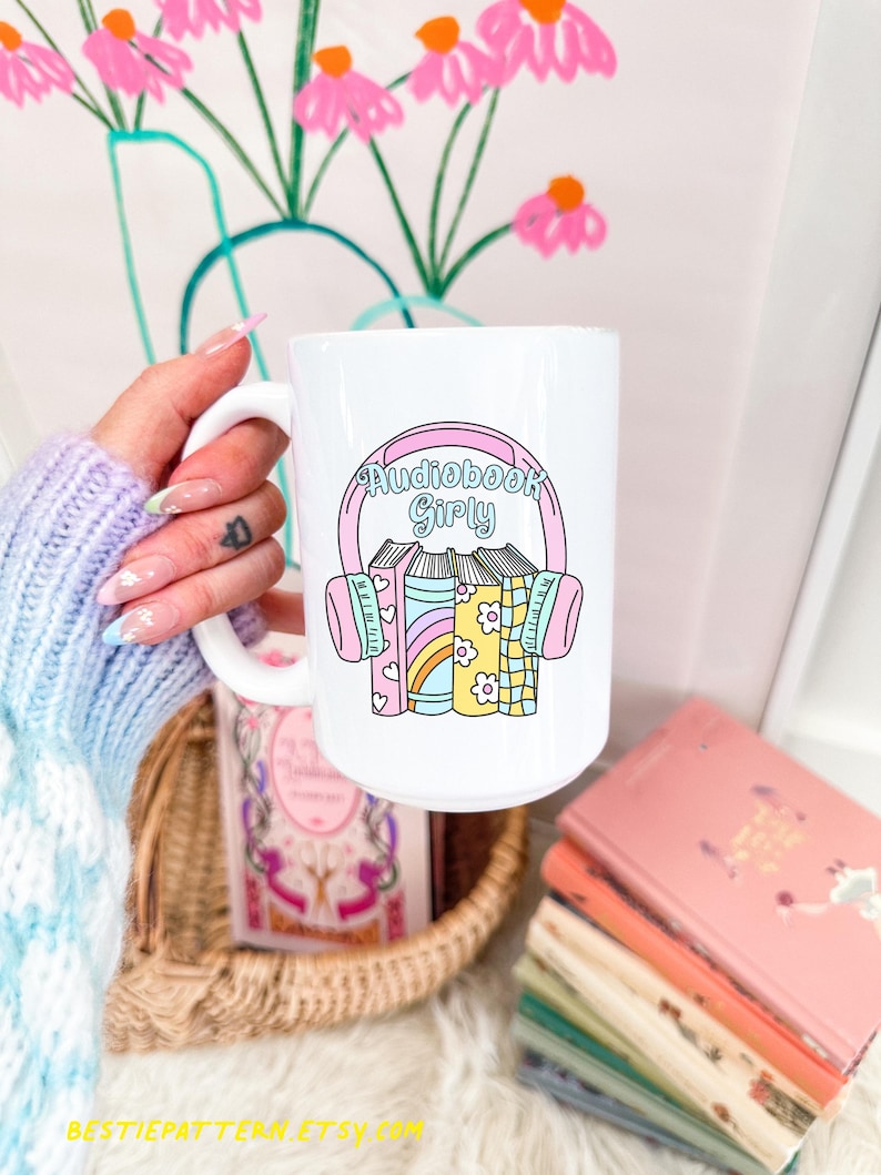 以下が含まれることがあります： ピンクと青のデザインで「Audiobook Girly」の文字が入った白いセラミックマグ。マグには、パステルカラーのヘッドフォンと本のグラフィックが描かれています。背景には本の山とバスケットがあります。