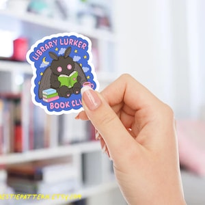 May include: A sticker featuring a cartoon moth reading a book, with the text "LIBRARY LURKER BOOK CLUB". The moth is black with pink eyes and is holding a green book. A pink mug and a stack of books are also visible.