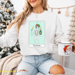May include: White sweatshirt with a green, white, and black illustration of a skeleton wearing a Santa hat and holding a glass of champagne and a bottle of champagne. The illustration is on a light blue background with the text "The Drinker" below it.