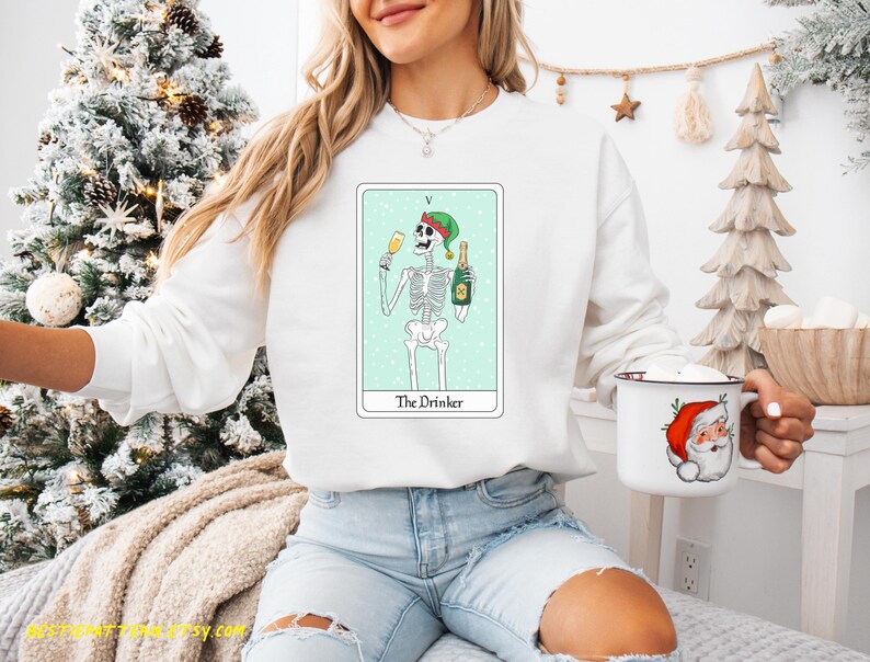 May include: White sweatshirt with a green, white, and black illustration of a skeleton wearing a Santa hat and holding a glass of champagne and a bottle of champagne. The illustration is on a light blue background with the text "The Drinker" below it.
