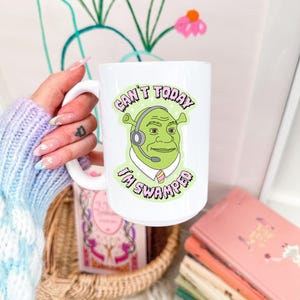 May include: White ceramic mug featuring a cartoon image of Shrek wearing a headset, with the text "Can't Today I'm Swamped." The mug has a large handle and is held by a person. The background includes a vase of pink flowers and a woven basket.