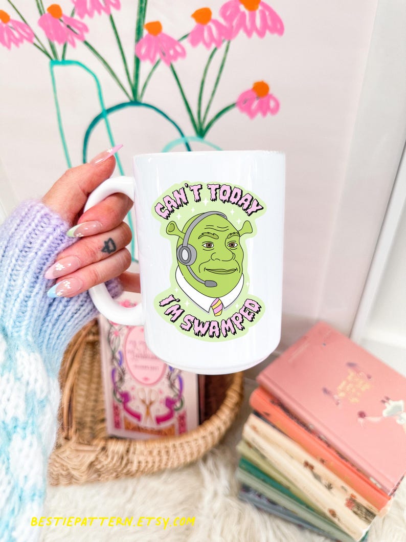 May include: White ceramic mug featuring a cartoon image of Shrek wearing a headset, with the text "Can't Today I'm Swamped." The mug has a large handle and is held by a person. The background includes a vase of pink flowers and a woven basket.