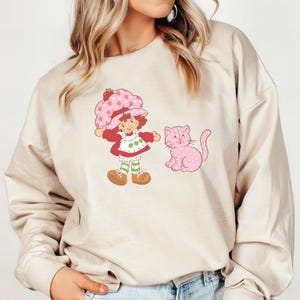 Puede incluir: Una sudadera de color crema con un estampado gr&aacute;fico de Strawberry Shortcake y un gato rosa. Strawberry Shortcake lleva un vestido rosa y verde y un sombrero rosa con una fresa en la parte superior. El gato est&aacute; sentado en el suelo y mirando hacia la derecha.