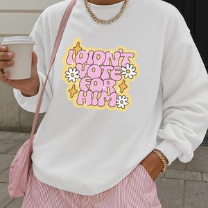 以下が含まれることがあります： ピンクのバブルレターで「I DIDN'T VOTE FOR HIM」と書かれた白いスウェットシャツ。黄色い星と白いデイジーに囲まれています。スウェットシャツは、ピンクと白のストライプのショーツと組み合わせています。