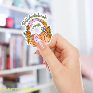 May include: A sticker featuring a pair of headphones with a pink and orange color scheme, adorned with fall leaves. The text "Fall Audiobook Girlie" is written above the headphones in a whimsical font. The sticker is held in a hand.