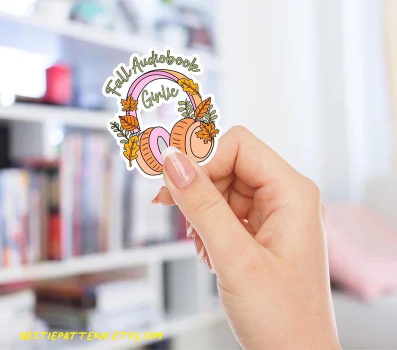 May include: A sticker featuring a pair of headphones with a pink and orange color scheme, adorned with fall leaves. The text "Fall Audiobook Girlie" is written above the headphones in a whimsical font. The sticker is held in a hand.