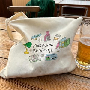 May include: A cream-colored tote bag with a variety of colorful illustrations, including a green desk lamp, books, and a coffee cup. The text "Meet me at the library" is printed on the bag. A glass of beer sits next to the bag.
