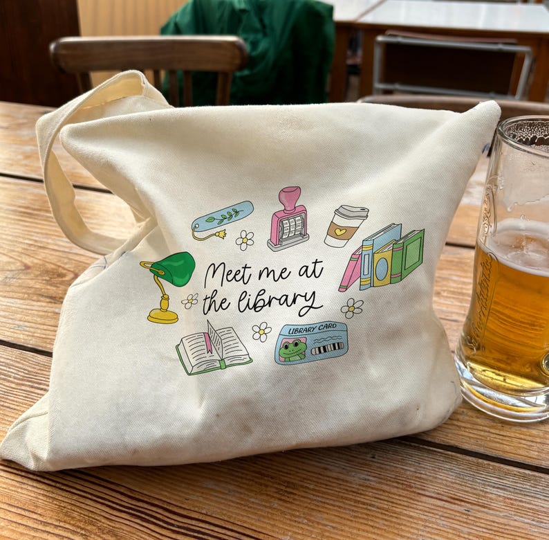 May include: A cream-colored tote bag with a variety of colorful illustrations, including a green desk lamp, books, and a coffee cup. The text "Meet me at the library" is printed on the bag. A glass of beer sits next to the bag.