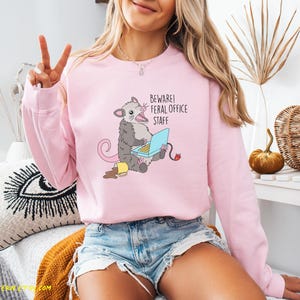 Puede incluir: Sudadera rosa claro con un dise&ntilde;o de zarig&uuml;eya de dibujos animados. La zarig&uuml;eya est&aacute; sentada y usando una computadora port&aacute;til, con el texto "BEWARE! FERAL OFFICE STAFF" encima. La sudadera tiene mangas largas.