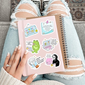 May include: A pink notebook with several colorful stickers, including "OUT OF OFFICE!" and "I RUN ON COFFEE, OFFICE GOSSIP AND SNARK." A brown spiral notebook is next to it. The person is wearing ripped blue jeans and white sneakers.