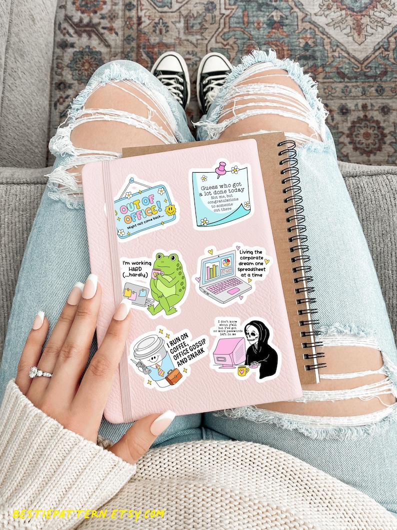 May include: A pink notebook with several colorful stickers, including "OUT OF OFFICE!" and "I RUN ON COFFEE, OFFICE GOSSIP AND SNARK." A brown spiral notebook is next to it. The person is wearing ripped blue jeans and white sneakers.
