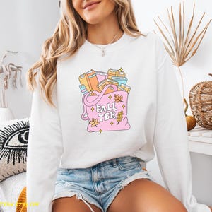 May include: White sweatshirt with a pink tote bag graphic filled with books. The tote bag has the text "FALL TBR" and autumn leaf accents. The sweatshirt is a long-sleeved, crew neck style.