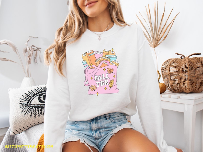 May include: White sweatshirt with a pink tote bag graphic filled with books. The tote bag has the text "FALL TBR" and autumn leaf accents. The sweatshirt is a long-sleeved, crew neck style.