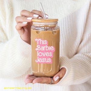 Puede incluir: Un vaso de vidrio transparente con tapa de bamb&uacute; y una pajita. El vaso est&aacute; lleno de caf&eacute; helado y tiene una pegatina rosa que dice "This Barbie loves Jesus".