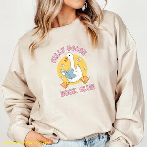 May include: Beige sweatshirt with the text "Silly Goose Book Club" in pink. The graphic features a white goose reading a blue book inside a yellow circle with small white flowers.