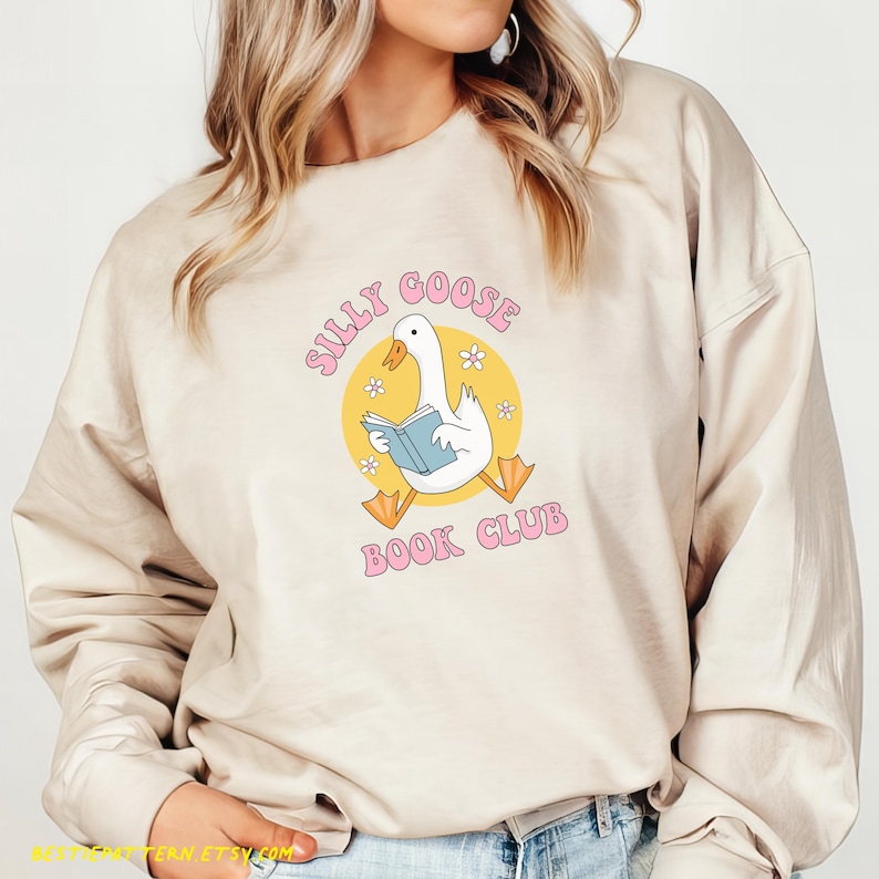 May include: Beige sweatshirt with the text "Silly Goose Book Club" in pink. The graphic features a white goose reading a blue book inside a yellow circle with small white flowers.