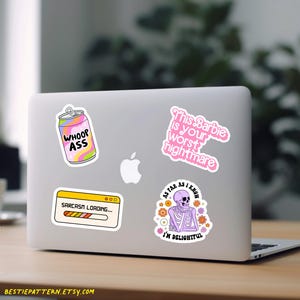 May include: Four stickers on a silver laptop. The stickers are: a pink can with the text "WHOOP ASS", a pink and purple skeleton with the text "As far as I know I'm delightful", a pink and white sticker with the text "This Barbie is your worst nightmare", and a yellow and orange progress bar with the text "Sarcasm Loading..."