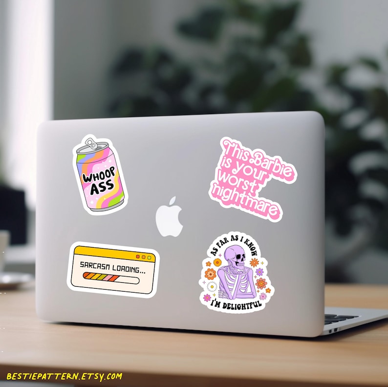 May include: Four stickers on a silver laptop. The stickers are: a pink can with the text "WHOOP ASS", a pink and purple skeleton with the text "As far as I know I'm delightful", a pink and white sticker with the text "This Barbie is your worst nightmare", and a yellow and orange progress bar with the text "Sarcasm Loading..."