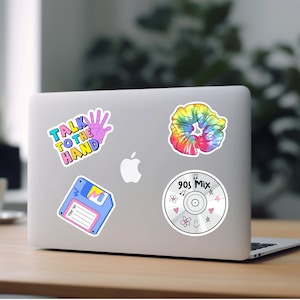 May include: A laptop computer with five stickers on it. The stickers are a floppy disk, a scrunchie, a hand with the text "Talk to the Hand", a CD with the text "90s Mix", and a pink heart.