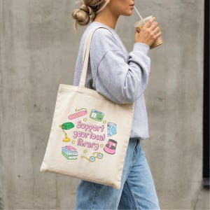 May include: Beige tote bag with the text "Support your local library" and colorful illustrations of books, glasses, a lamp, and other library-themed items. The bag is carried over the shoulder.