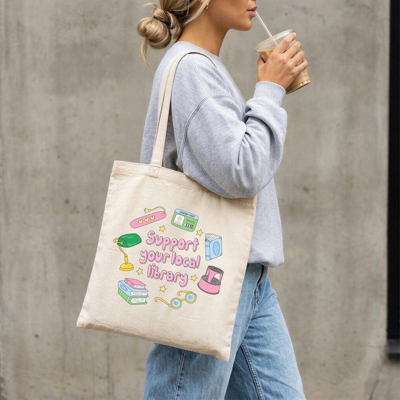 May include: Beige tote bag with the text "Support your local library" and colorful illustrations of books, glasses, a lamp, and other library-themed items. The bag is carried over the shoulder.