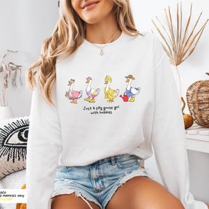 May include: White sweatshirt with a graphic of four geese wearing different outfits and the text "Just a silly goose girl with hobbies".