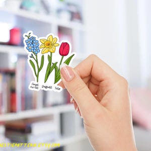 以下が含まれることがあります： 青いワスレナグサ、黄色のスイセン、赤いチューリップの3つの花が描かれたステッカー。ステッカーには「Forget me not, Daffodil, Tulip」の文字が入っています。ステッカーは手で持たれています。