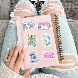 May include: A pink notebook adorned with library-themed stickers, including a frog with a library card and a goose with books. A spiral-bound notebook rests beside it. The person is wearing ripped jeans and black and white sneakers.