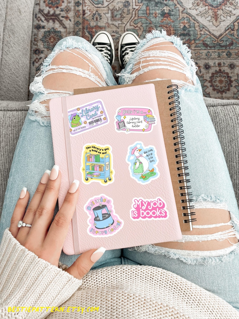 May include: A pink notebook adorned with library-themed stickers, including a frog with a library card and a goose with books. A spiral-bound notebook rests beside it. The person is wearing ripped jeans and black and white sneakers.
