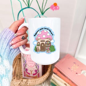 May include: White ceramic mug featuring a whimsical mushroom-shaped library design with the text "Support your local library." The mug has a large handle and is surrounded by books and a basket.