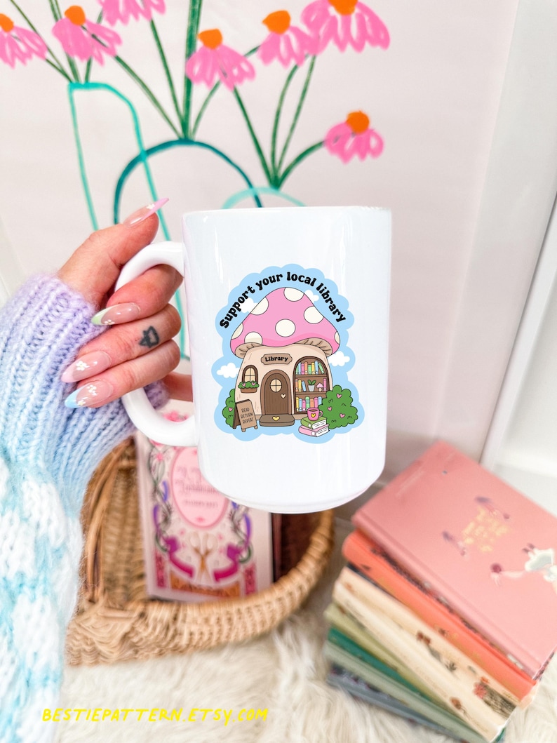 May include: White ceramic mug featuring a whimsical mushroom-shaped library design with the text "Support your local library." The mug has a large handle and is surrounded by books and a basket.