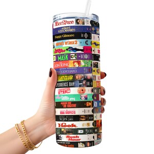 May include: A stainless steel tumbler with a colorful design featuring the titles of popular movies from the 1990s. The tumbler is wrapped with VHS tape covers, including "Office Space", "Edward Scissorhands", "Happy Gilmore", "The Lion King", "The Bodyguard", "Independence Day", "American Pie", "The Fugitive", "Hook", and "Rush Hour".