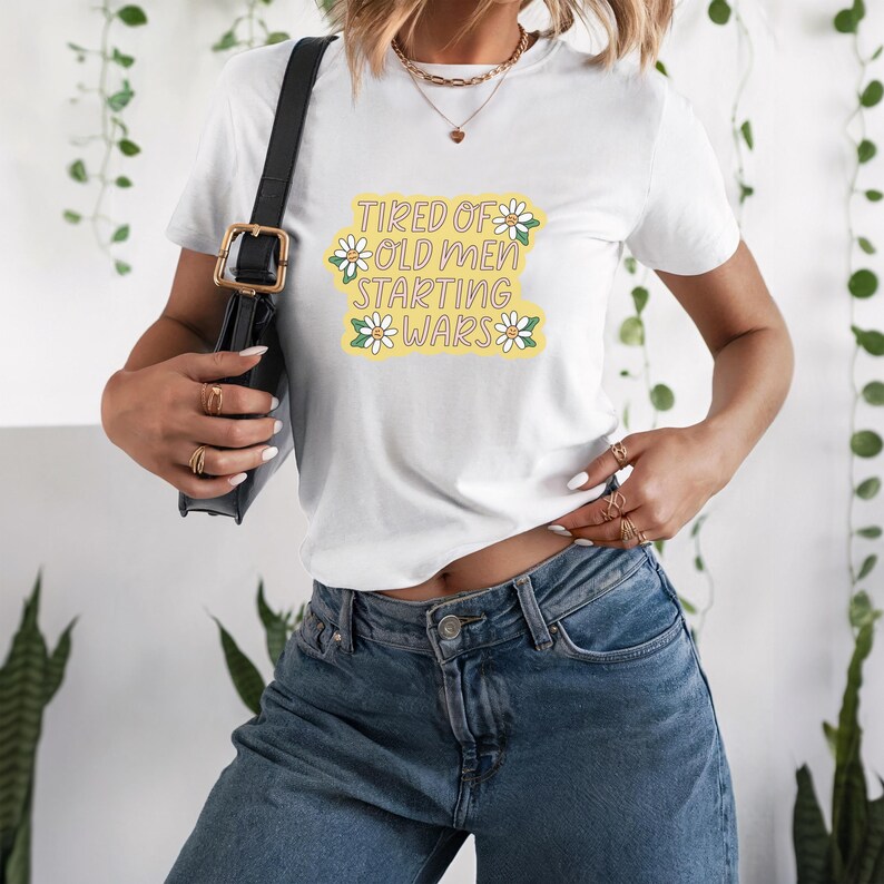 以下が含まれることがあります： 「Tired of Old Men Starting Wars」の文字が黄緑色の花柄でプリントされた白いTシャツ。青いジーンズと黒いショルダーバッグと合わせています。