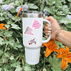 以下が含まれることがあります： シルバーの縁、ハンドル、蓋が付いた白い断熱タンブラー。タンブラーには、テキスト「No more fucking wars」のステッカー1枚、セージバンドルとテキスト「Not enough sage for this administration」のステッカー1枚、オポッサムとテキスト「My tummy hurts and Trump is bombing the Middle Earth」のステッカー1枚が付いています。