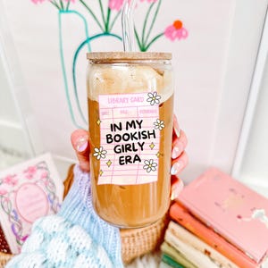 以下が含まれることがあります： 木製の蓋とガラスのストローが付いた透明なガラスのタンブラーで、コーヒー飲料が入っています。「IN MY BOOKISH GIRLY ERA」と書かれたステッカーと花のアクセントが付いています。背景には本と花のプリントがあります。