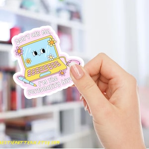 May include: A sticker featuring a cartoon laptop with a smiling face on the screen and floral accents. The text on the sticker reads "Don't ask me, I'm the personality hire." A pen and mouse are also included in the design. The sticker is held by a hand.