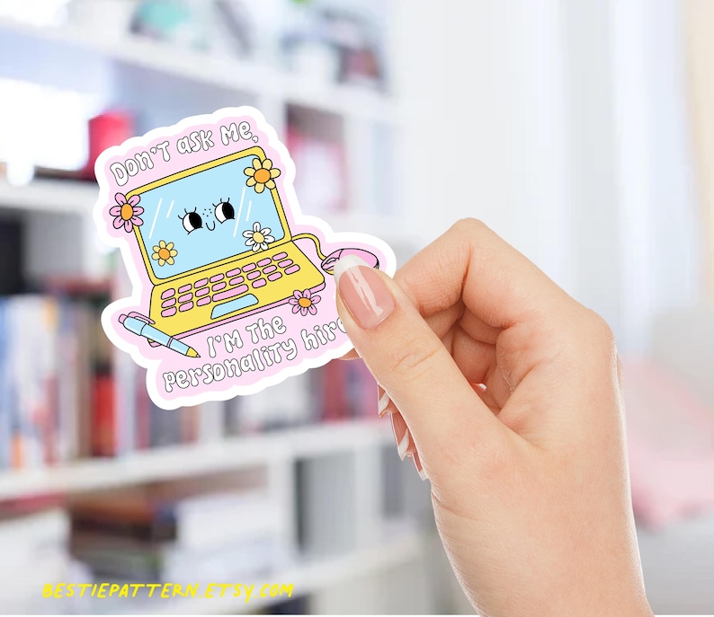 May include: A sticker featuring a cartoon laptop with a smiling face on the screen and floral accents. The text on the sticker reads "Don't ask me, I'm the personality hire." A pen and mouse are also included in the design. The sticker is held by a hand.
