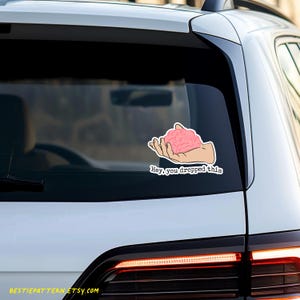 May include: A pink brain in a hand with the text "Hey, you dropped this" on a white car.