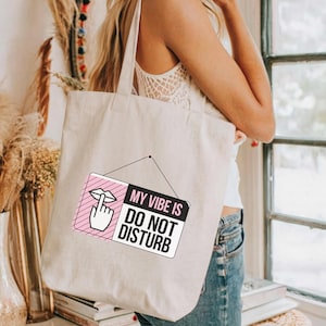 May include: A white tote bag with a pink and black sign that says "My Vibe Is Do Not Disturb". The sign has a pink lips and finger emoji on it.