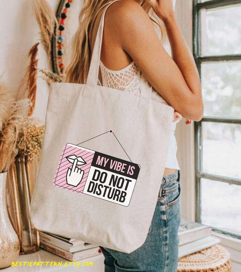 May include: A white tote bag with a pink and black sign that says "My Vibe Is Do Not Disturb". The sign has a pink lips and finger emoji on it.