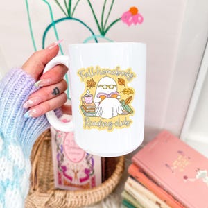 May include: White ceramic mug with a wraparound graphic featuring a ghost reading a book, with the text "Fall homebody Reading club". The mug has a large handle and is filled with a beverage. A stack of books is visible.
