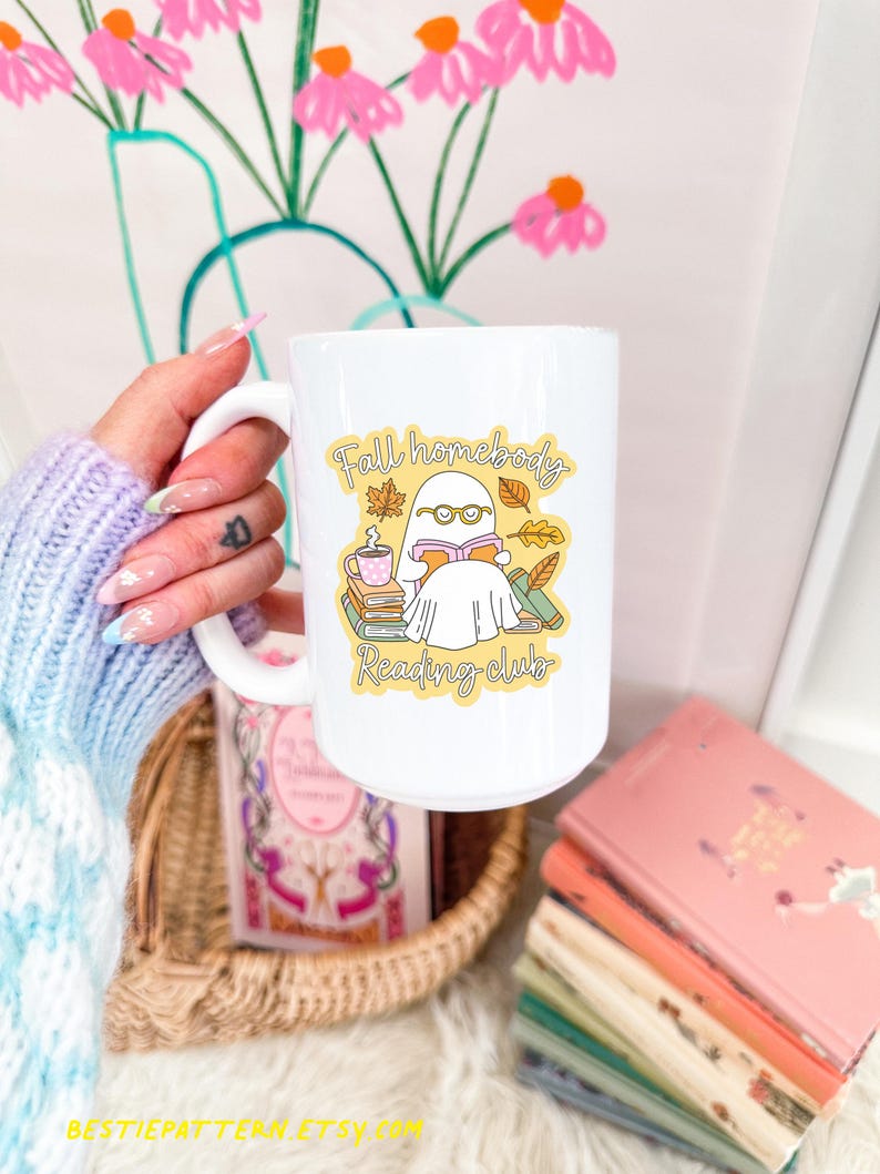 May include: White ceramic mug with a wraparound graphic featuring a ghost reading a book, with the text "Fall homebody Reading club". The mug has a large handle and is filled with a beverage. A stack of books is visible.