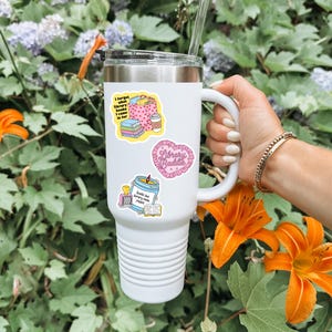 May include: White insulated tumbler with a black lid and a white handle. The tumbler is decorated with colorful stickers, including one that says "Library Baddie" in a heart shape. The background features orange lilies and green foliage.