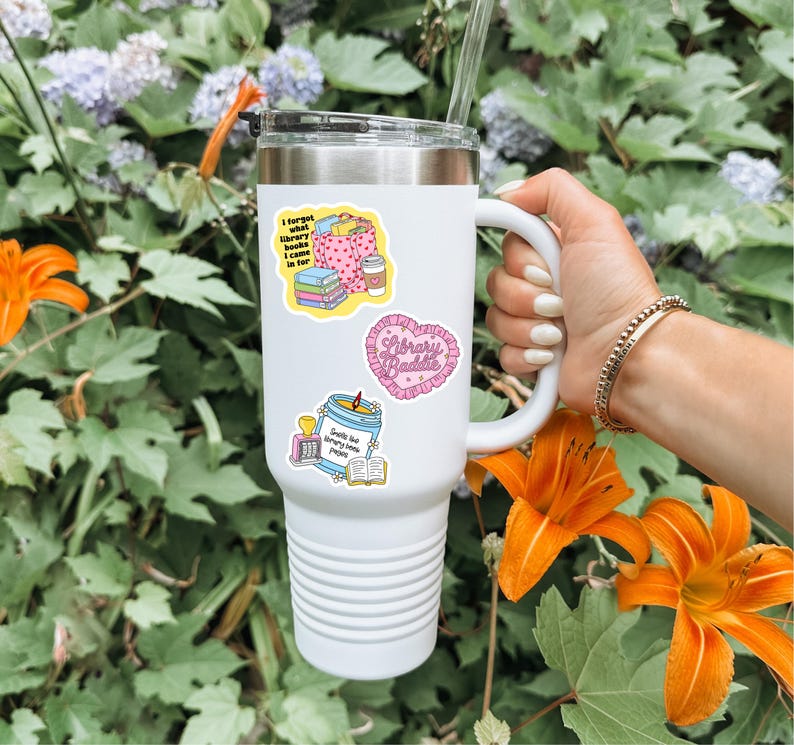 May include: White insulated tumbler with a black lid and a white handle. The tumbler is decorated with colorful stickers, including one that says "Library Baddie" in a heart shape. The background features orange lilies and green foliage.