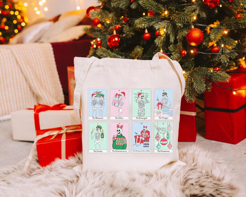 May include: A white canvas tote bag with a colorful design of skeletons celebrating Christmas. The bag features the text "The Misletoe", "The Office", "The Reindeer", "The Christmas Sweater", "The Christmas Movie Marathon", and "The Ornaments".