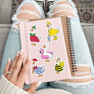May include: A set of six goose stickers featuring different costumes. The stickers are on a pink notebook with a spiral binding. The stickers include a goose wearing a strawberry costume, a goose wearing a raincoat, a goose wearing a life preserver, a goose wearing a beret and a striped shirt, and a goose wearing a bee costume.