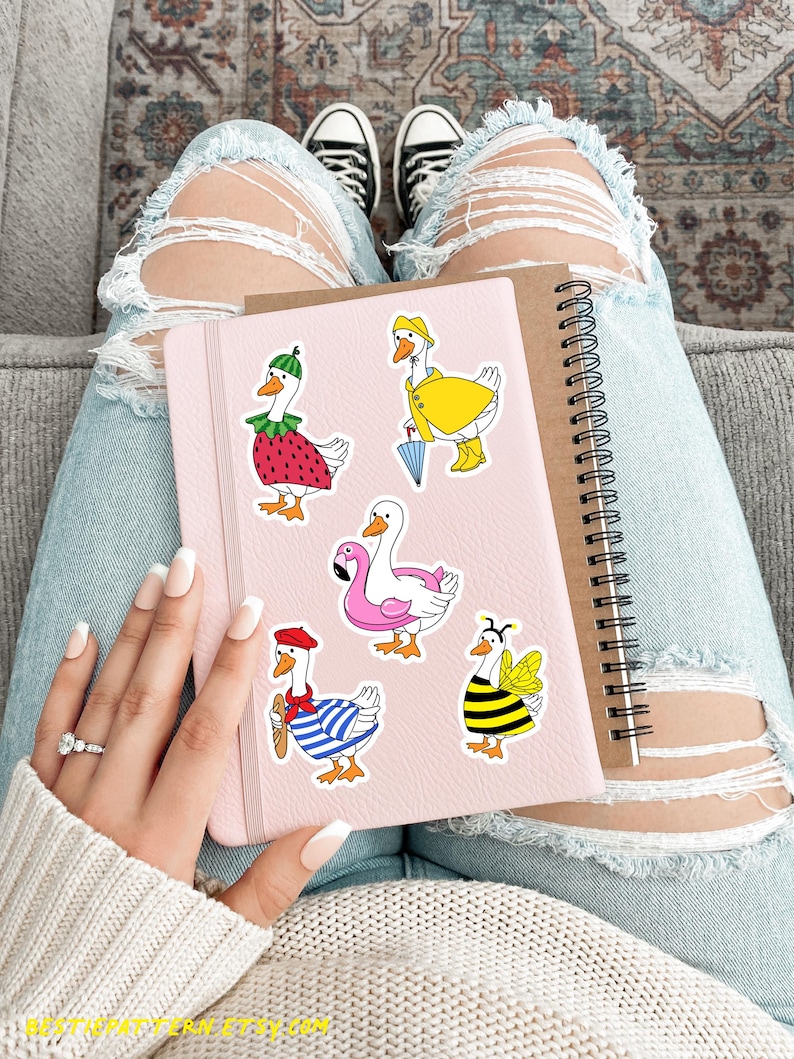 May include: A set of six goose stickers featuring different costumes. The stickers are on a pink notebook with a spiral binding. The stickers include a goose wearing a strawberry costume, a goose wearing a raincoat, a goose wearing a life preserver, a goose wearing a beret and a striped shirt, and a goose wearing a bee costume.