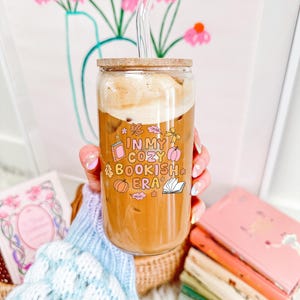 May include: A clear glass tumbler with a wooden lid and a glass straw, filled with a coffee drink. The tumbler has the text "IN MY COZY BOOKISH ERA" with book and pumpkin graphics. The tumbler is held in front of a floral background.