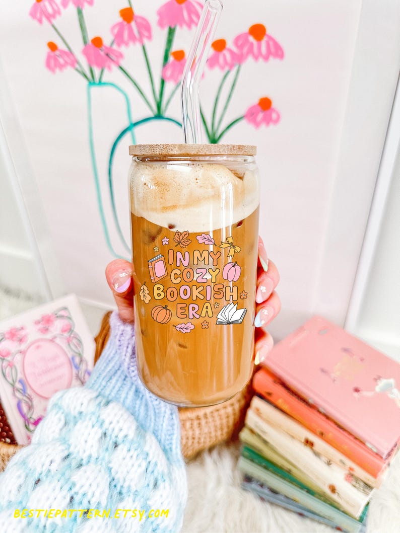 May include: A clear glass tumbler with a wooden lid and a glass straw, filled with a coffee drink. The tumbler has the text "IN MY COZY BOOKISH ERA" with book and pumpkin graphics. The tumbler is held in front of a floral background.